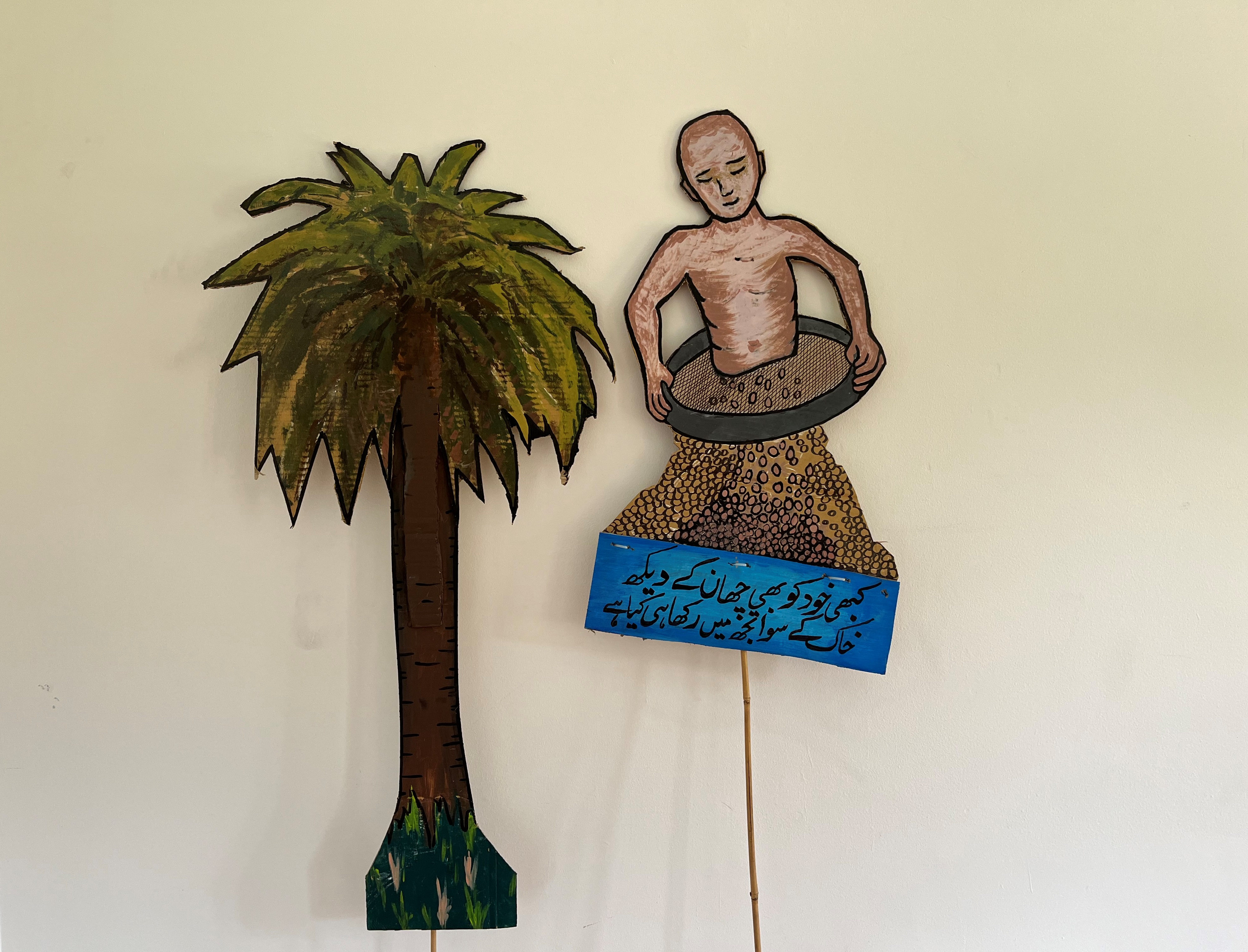 A bald person sits bare chested on what looks like a round sieve small rocks are dropping and have created a mound, at the bottom of the mound is a blue plaque with calligraphy type writing in either farsi or arabic